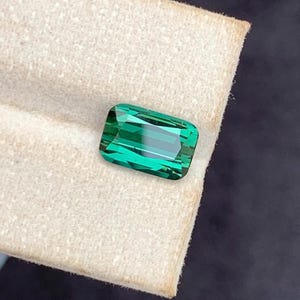 May include: A rectangular, emerald-cut gemstone in a vivid green colour. The gem is displayed on a cream-coloured fabric, with a dark background. The facets of the stone reflect light, creating a sparkling effect.