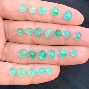 May include: A selection of small, polished gemstones in various shapes, including oval, round, and square. The gems display a range of green and teal hues, with a translucent quality. The stones are displayed on a hand.