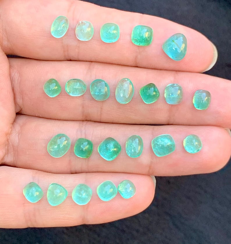 May include: A selection of small, polished gemstones in various shapes, including oval, round, and square. The gems display a range of green and teal hues, with a translucent quality. The stones are displayed on a hand.