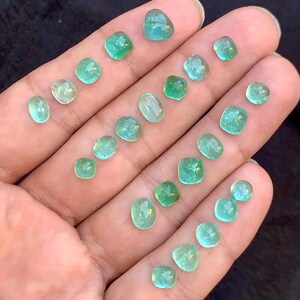 May include: A selection of small, polished emerald gemstones in various shapes, including round, oval, and square. The gems are a vibrant green colour and are displayed on a hand, showcasing their clarity and brilliance.