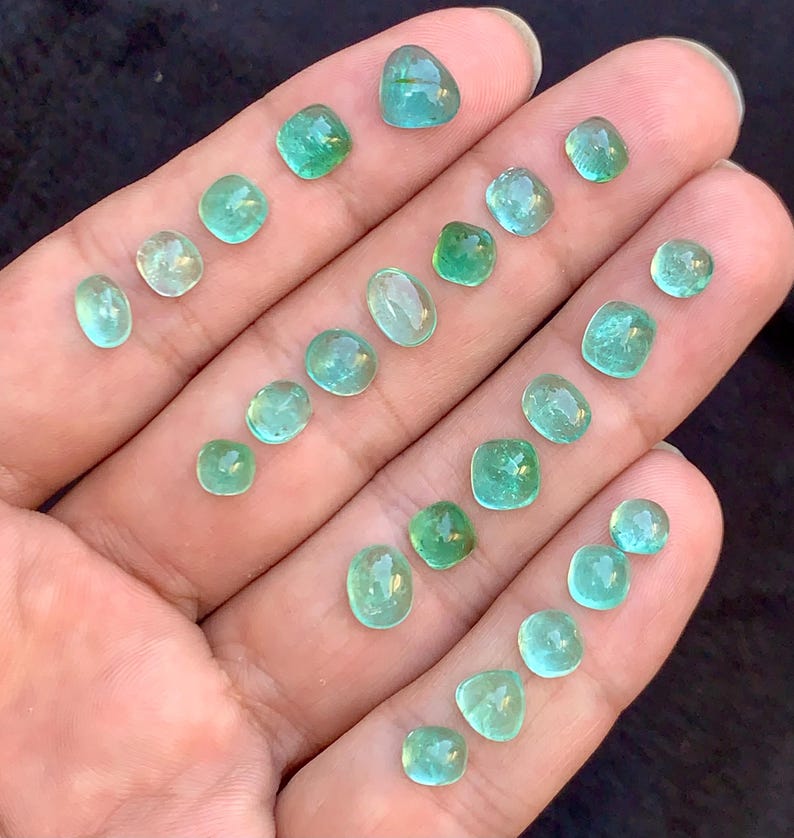 May include: A selection of small, polished emerald gemstones in various shapes, including round, oval, and square. The gems are a vibrant green colour and are displayed on a hand, showcasing their clarity and brilliance.