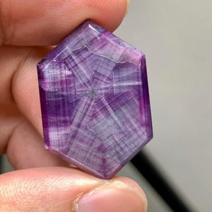 May include: A hexagonal gemstone, polished to a high sheen, displaying a rich purple colour. The stone's interior reveals a complex pattern of intersecting lines and subtle colour variations. The gemstone is held between fingers.