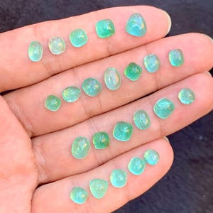 May include: A hand holds a selection of small, polished gemstones in shades of teal and green. The stones are in various shapes, from rounded to square, and have a translucent appearance. The gems are smooth and reflect light.