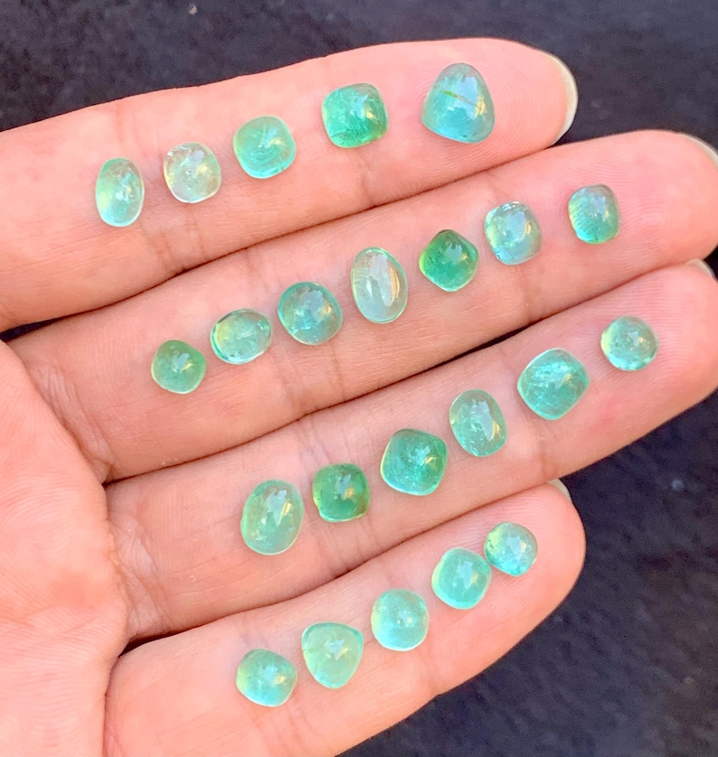 May include: A hand holds a selection of small, polished gemstones in shades of teal and green. The stones are in various shapes, from rounded to square, and have a translucent appearance. The gems are smooth and reflect light.