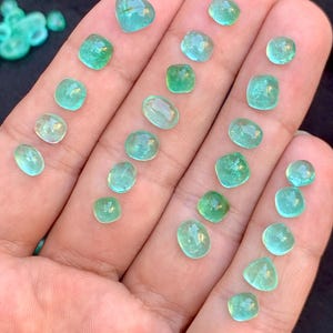 May include: A selection of small, polished, emerald-green gemstones in various shapes, such as oval, square, and teardrop, are displayed on a hand. The gems are translucent, with slight colour variations.