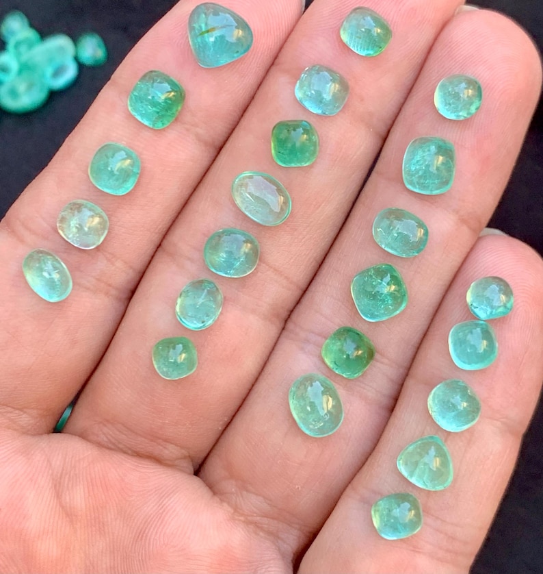 May include: A selection of small, polished, emerald-green gemstones in various shapes, such as oval, square, and teardrop, are displayed on a hand. The gems are translucent, with slight colour variations.