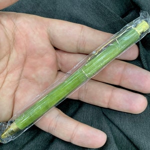 May include: A green jade pen with gold accents is wrapped in clear plastic and held in a hand.