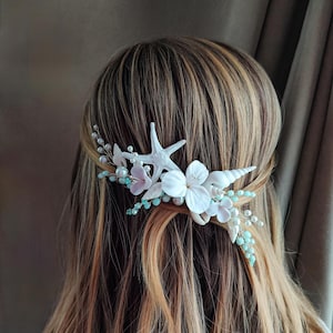 Op de afbeelding: Een haar kam met een witte zeester en schelp, witte en roze bloemen en blauwe kralen.