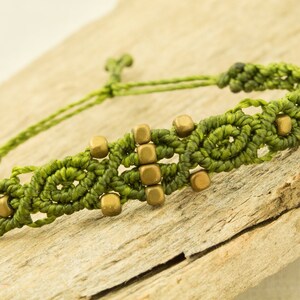 Könnte beinhalten: Ein grünes Makramee-Armband mit goldenen quadratischen Perlen. Das Armband ist mit einem Knoten gebunden und liegt auf einem Stück Holz.