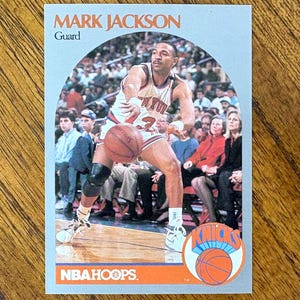May include: A Mark Jackson basketball card from NBA Hoops. The card features a photo of Jackson in a New York Knicks uniform, dribbling a basketball. The card has the Knicks logo and the NBA Hoops logo.