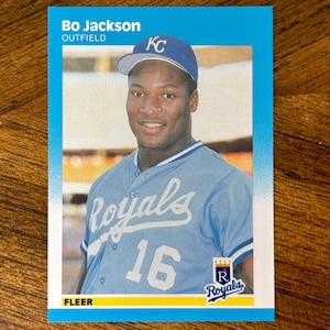 May include: A baseball card featuring Bo Jackson in a Kansas City Royals uniform. The card has a blue border and includes the text "Bo Jackson OUTFIELD" at the top. The player is wearing a blue cap and jersey with the number 16.