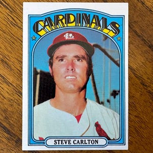 May include: A vintage baseball card featuring Steve Carlton. The card has a blue border with the word "CARDINALS" at the top. Carlton is wearing a red cap and a white jersey. The text "STEVE CARLTON" is at the bottom.