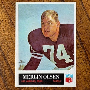 May include: Vintage football card featuring Merlin Olsen of the Los Angeles Rams. The card shows Olsen in his purple and white uniform, with the number 74 prominently displayed. The NFL logo is visible.