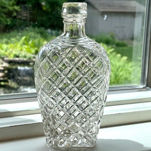 May include: A clear glass bottle with a diamond pattern. The bottle has a round body and a tall neck.