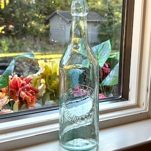 May include: A clear, antique glass bottle with a textured surface. The bottle has a long neck and a rounded body. The bottle is positioned in front of a window with a view of greenery and flowers.