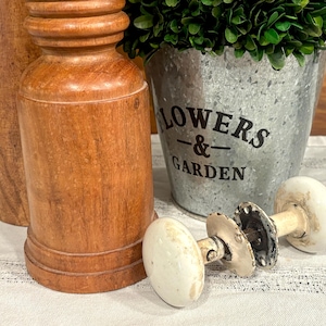 May include: A rustic still life featuring wooden decorative items and a galvanized metal bucket with the words "FLOWERS & GARDEN". Two antique white porcelain door knobs are in the foreground.