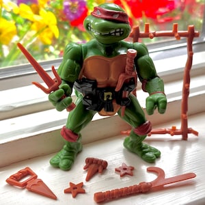 May include: A green Teenage Mutant Ninja Turtle action figure with a red bandana, holding a sai. The figure has a brown shell and black belt. Additional orange weapons, including stars and a hook, are placed in front of the figure.