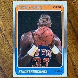 May include: A Fleer trading card featuring Patrick Ewing of the New York Knicks. The card has an orange top border with the player's name, and a blue bottom border with "Knickerbockers" and "Center". Ewing is holding a basketball.