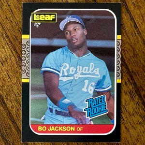 May include: A baseball card featuring Bo Jackson of the Kansas City Royals. The card has a black border with a yellow and white design on the left side. Jackson is wearing a light blue Royals jersey with the number 16. The card also has a "Rated Rookie" emblem.