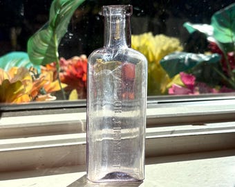 May include: A clear, rectangular antique bottle with the text "E. OR. J. INIMENT" embossed vertically. The bottle is slightly tinted and sits on a windowsill with a blurred background of colorful flowers and foliage.