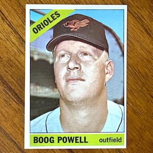 May include: A vintage baseball card depicting Boog Powell of the Orioles. The card shows Powell wearing a black cap with the Orioles logo. The word "Orioles" is in a green banner at the top.