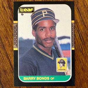 May include: A baseball trading card featuring Barry Bonds of the Pittsburgh Pirates. The card has a black border with a green bottom and the text "BARRY BONDS OF". The image shows Bonds wearing a Pirates cap and uniform, holding a baseball bat.