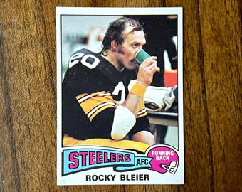 May include: A vintage football trading card featuring Rocky Bleier of the Pittsburgh Steelers. The card shows Bleier drinking from a green cup, wearing his black and gold Steelers uniform. The card has the Steelers logo and the words "Running Back".