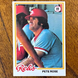 May include: A baseball card featuring Pete Rose of the Cincinnati Reds. The card has a white border and a photo of Rose in a red helmet and light blue jersey. The Reds logo is at the bottom left, and "PETE ROSE" is printed below the image.