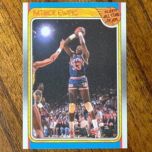 May include: A Patrick Ewing basketball card featuring a player in a blue and orange uniform, mid-jump, shooting a basketball. The card has a blue border and the text "PATRICK EWING" and "FLEER ALL STAR TEAM".