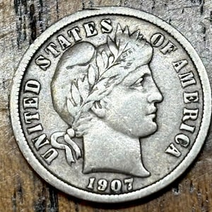 May include: A silver 1907 United States of America coin. The coin features a profile of a woman with a laurel wreath and the text "UNITED STATES OF AMERICA" around the edge. The year "1907" is visible at the bottom.