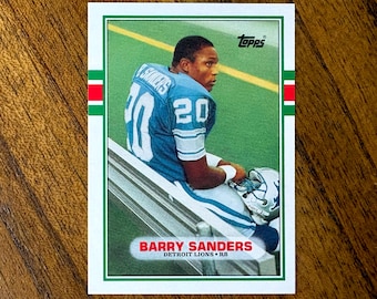 Può includere: Una figurina Topps con Barry Sanders dei Detroit Lions. Sanders indossa una maglia blu e bianca con il numero 20. La figurina ha un bordo verde e bianco con il logo Topps.