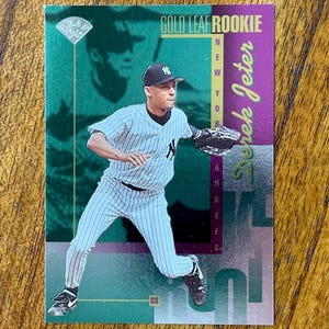 May include: A baseball card featuring Derek Jeter in a New York Yankees uniform. The card has a green and purple background with the text "Gold Leaf Rookie." Jeter is shown in a fielding pose.