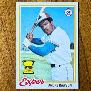 May include: A vintage baseball card featuring Andre Dawson of the Expos. The card has a yellow border and displays a color image of Dawson in his uniform, holding a bat. The card also includes the Expos logo and the text "Andre Dawson".
