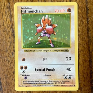May include: A Hitmonchan Pokemon card from the 1999 Wizards of the Coast set. The card is yellow and features an illustration of Hitmonchan, a fighting-type Pokemon, with a green background. The card text reads "Basic Pokemon Hitmonchan 70 HP Punching Pokemon. Length: 4' 7", Weight: 111 lbs. Jab 20 Special Punch 40 Weakness Resistance Retreat Cost While seeming to do nothing, it fires punches in lightning-fast volleys that are impossible to see. LV.33 #107 Illus. Ken Sugimori 1995, 96, 98, 99 Nintendo Creatures GAMEFREAK 1999 Wizards 7/102"