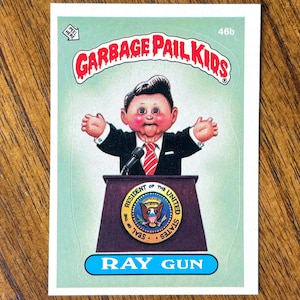 May include: A Garbage Pail Kids trading card featuring a cartoon character in a suit and tie behind a podium with the presidential seal. The card is labeled "Ray Gun" in a blue banner.