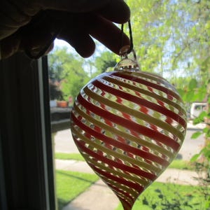 May include: A teardrop-shaped glass ornament featuring a clear base with swirling red and yellow stripes. The ornament has a small glass loop at the top for hanging and is suspended by a thin dark string, showcasing a translucent, spiraling design.