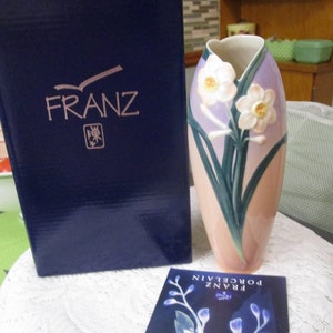 May include: A porcelain vase with a pink and purple gradient design featuring white daffodils with green leaves. The vase is in a blue box with the word "FRANZ" printed on it.