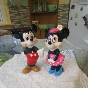 May include: A vintage ceramic figurine set of Mickey Mouse and Minnie Mouse. Mickey is wearing red shorts and yellow shoes. Minnie is wearing a pink dress with white polka dots and blue shoes.