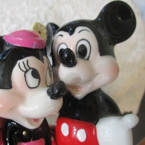 May include: A ceramic figurine of Mickey Mouse and Minnie Mouse. Mickey is wearing red shorts with white polka dots and Minnie is wearing a pink dress with a white flower.