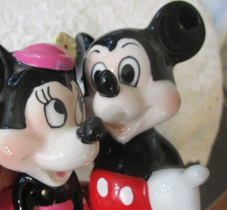 May include: A ceramic figurine of Mickey Mouse and Minnie Mouse. Mickey is wearing red shorts with white polka dots and Minnie is wearing a pink dress with a white flower.