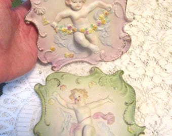 Puede incluir: Dos placas de pared de porcelana con querubines y marcos ornamentados. Los querubines están representados en una pose juguetona con detalles florales. Una placa tiene un fondo rosa y la otra un fondo verde.