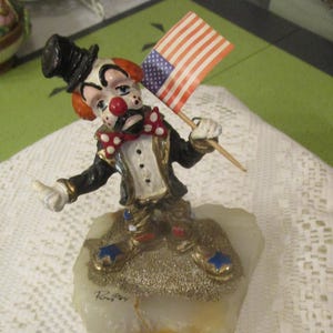 May include: A ceramic clown figurine holding a miniature American flag. The clown wears a black top hat, a red bow tie, and a black and gold tuxedo. The figurine stands on a white and gold base.