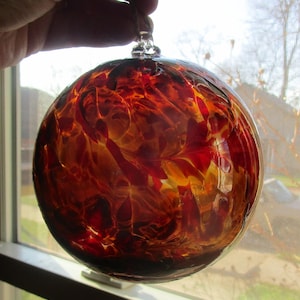 May include: A clear glass ornament with a fiery, abstract design in shades of red, orange, and yellow. The round ornament has a loop for hanging and is held up by a hand. The glass has a textured appearance.