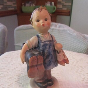 May include: A porcelain figurine of a young child holding a satchel and a pair of shoes. The child wears a blue denim apron over a white shirt. The figurine has rosy cheeks and is mounted on a round, off-white base.
