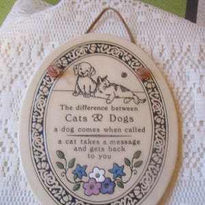 May include: Oval ceramic plaque with a dog and cat illustration, featuring the text "The difference between Cats & Dogs." The plaque has a decorative border, floral accents, and a leather hanging loop.