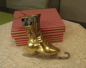 May include: A gold-colored ice skate ornament with decorative blue, red, and purple accents. The ornament has a silver blade and a gold loop for hanging. A red and white striped box is in the background.
