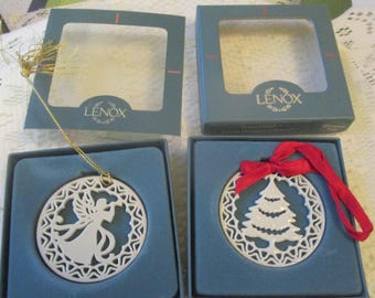 May include: Two white porcelain circular ornaments in blue Lenox boxes. One ornament features a cutout angel with a gold string, and the other features a cutout Christmas tree with a red ribbon. Both ornaments have intricate lattice borders.