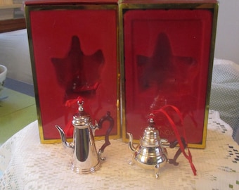 May include: Two silver-toned metal ornaments shaped like a coffee pot and a teapot, each featuring a dark brown handle and a red ribbon for hanging. They are displayed in front of two rectangular red boxes with gold-colored trim and molded interiors.