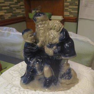 May include: A ceramic figurine depicting Santa Claus with two children. The figures are dressed in dark blue coats and hats, with Santa's beard and the children's faces in a light beige color. A sack of toys sits at Santa's feet.