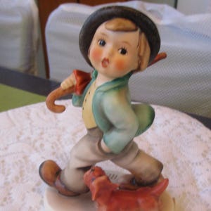 May include: A porcelain figurine of a boy wearing a blue jacket and a brown hat, holding an umbrella and walking with a brown dog.